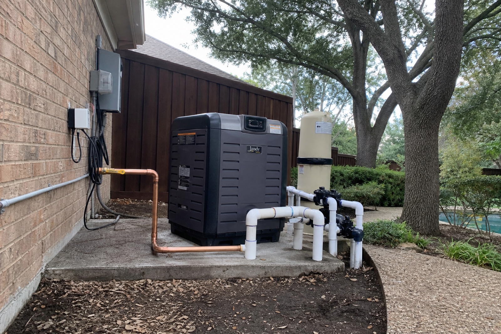 Newly installed natural gas pool heater on a concrete pad with fresh copper gas line and white PVC plumbing connections, oak tree overhead.