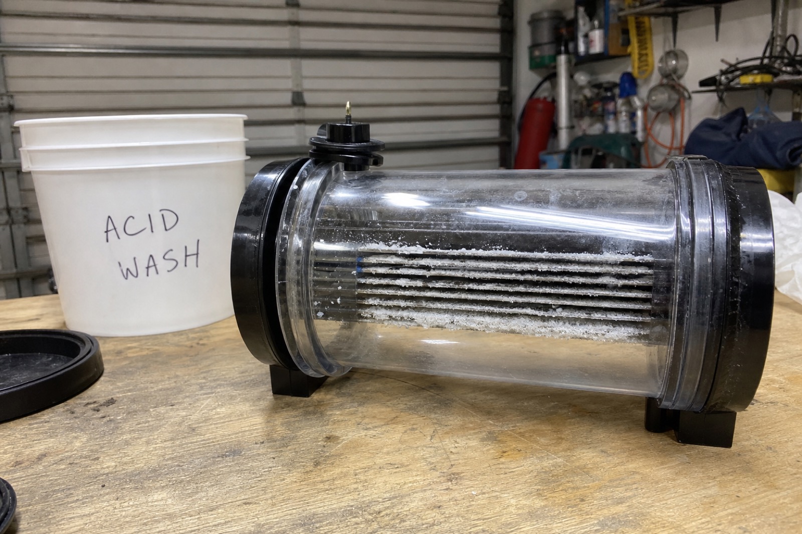 Pool salt cell on a workbench showing visible calcium scale buildup on the titanium plates, with an acid wash bucket beside it.