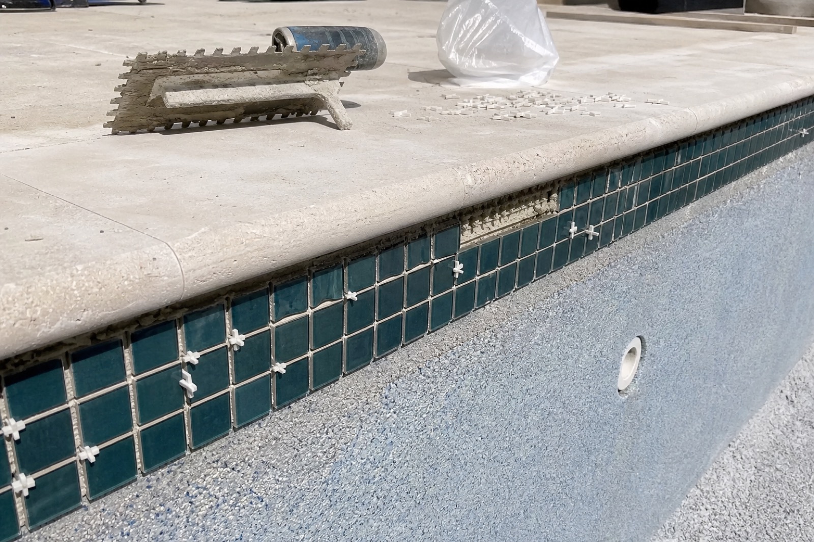A band of dark teal mosaic tile being installed at the waterline of a new Seahorse pool, with thinset trowel and spacers nearby.